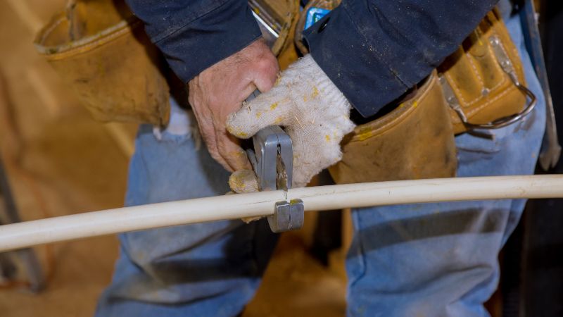 Gloved worker with a leather tool belt using a hand tool on a cream-colored plastic pipe