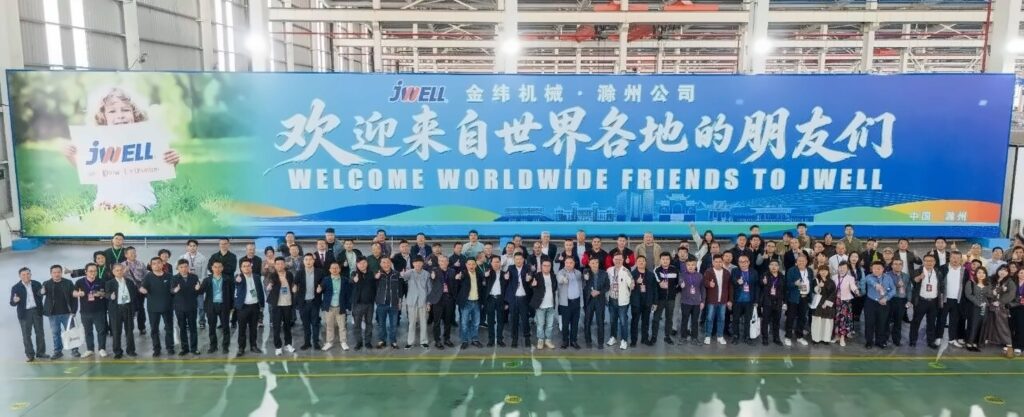Group photo of delegates on the JWELL factory floor under a Welcome Worldwide Friends to JWELL banner